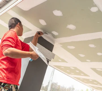 Gypsum ceiling installation in progress with a worker applying joint compound and finishing drywall panels on a suspended ceiling structure during interior construction 