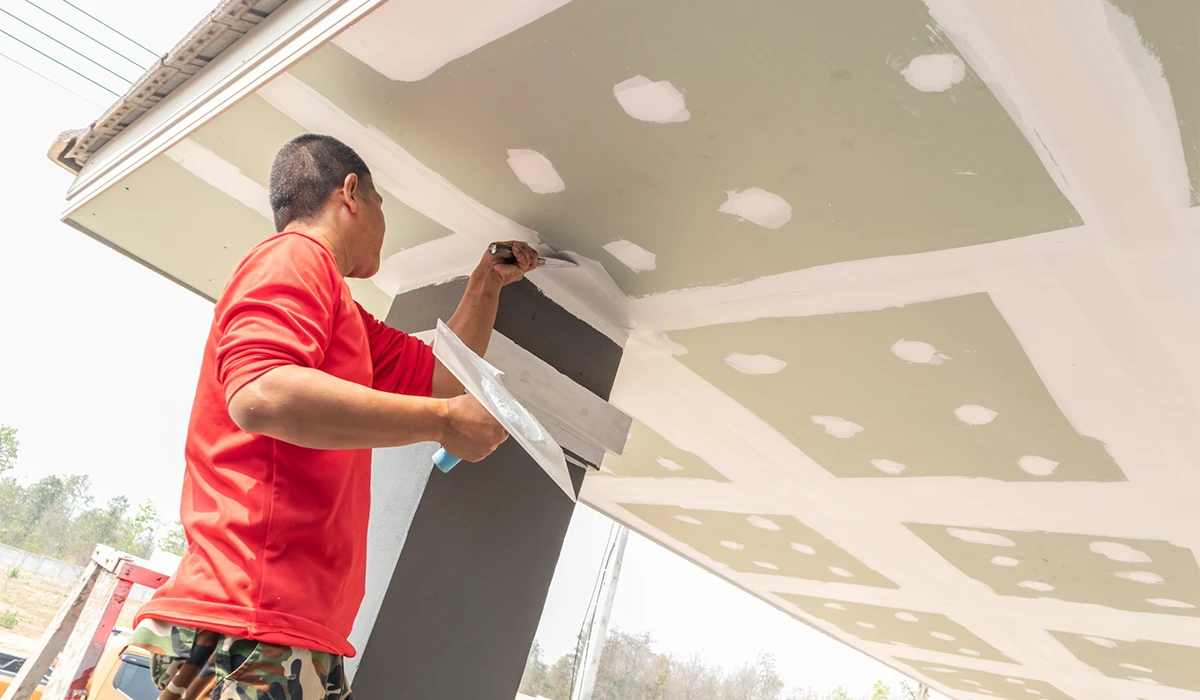 Lumens MEP Gypsum ceiling installation in progress with a worker applying joint compound and finishing drywall panels on a suspended ceiling structure during interior construction